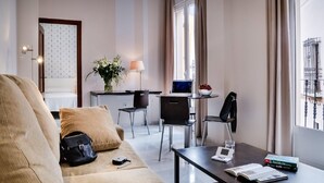 Una habitación de hotel moderna con sofá, una zona de comedor con mesa y sillas, un escritorio con computadora y una ventana con cortinas.