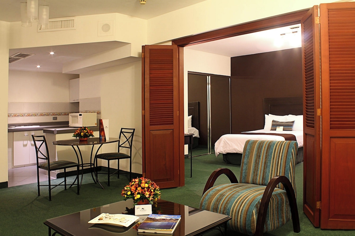 Habitación de hotel con cama, zona de comedor con mesa y sillas, una kitchenette pequeña y un baño visible a través de una puerta abierta.