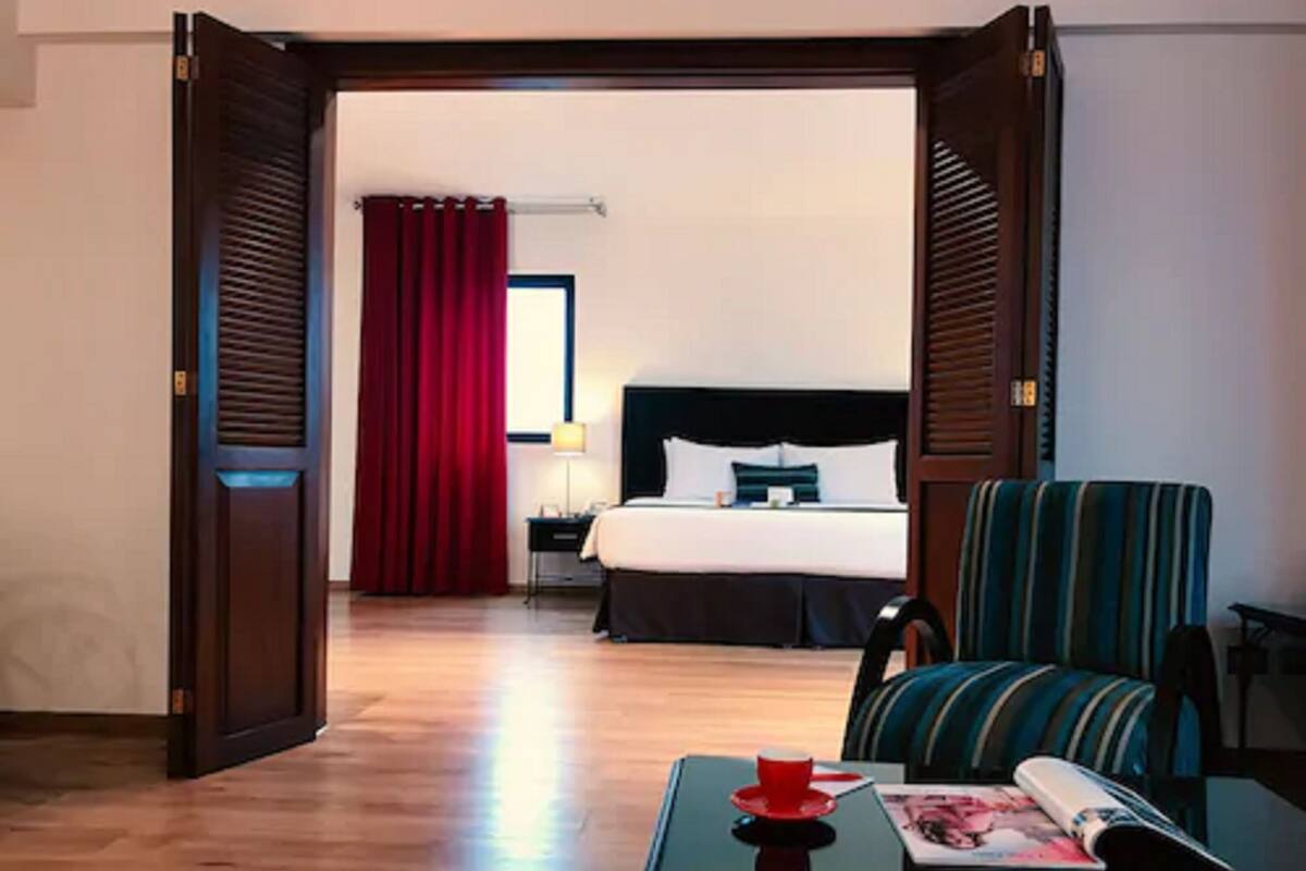 Une chambre d’hôtel avec un lit, une table de chevet, une lampe, un rideau rouge, un fauteuil à rayures et une table sur laquelle se trouvent une tasse rouge et un magazine.