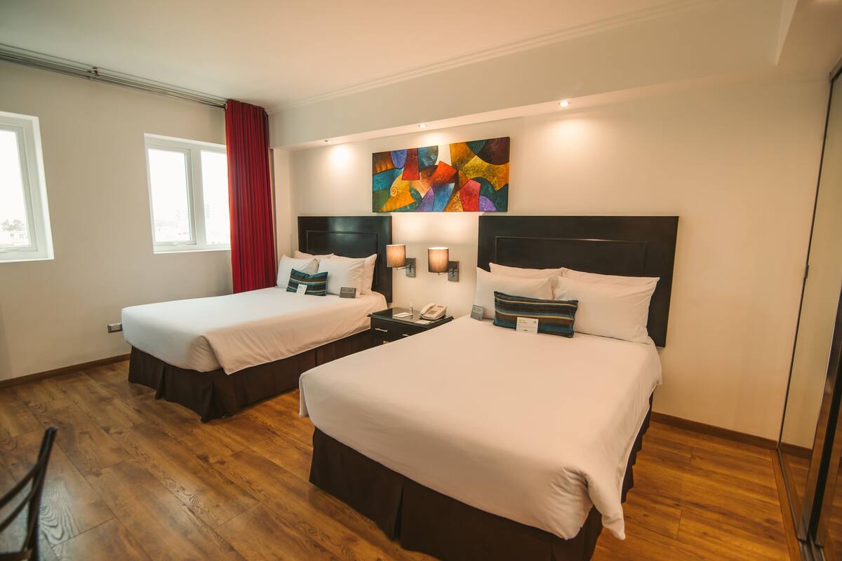 Une chambre d’hôtel avec deux lits, un plancher en bois, un rideau rouge, une peinture abstraite aux couleurs vives et une table de chevet avec une lampe.