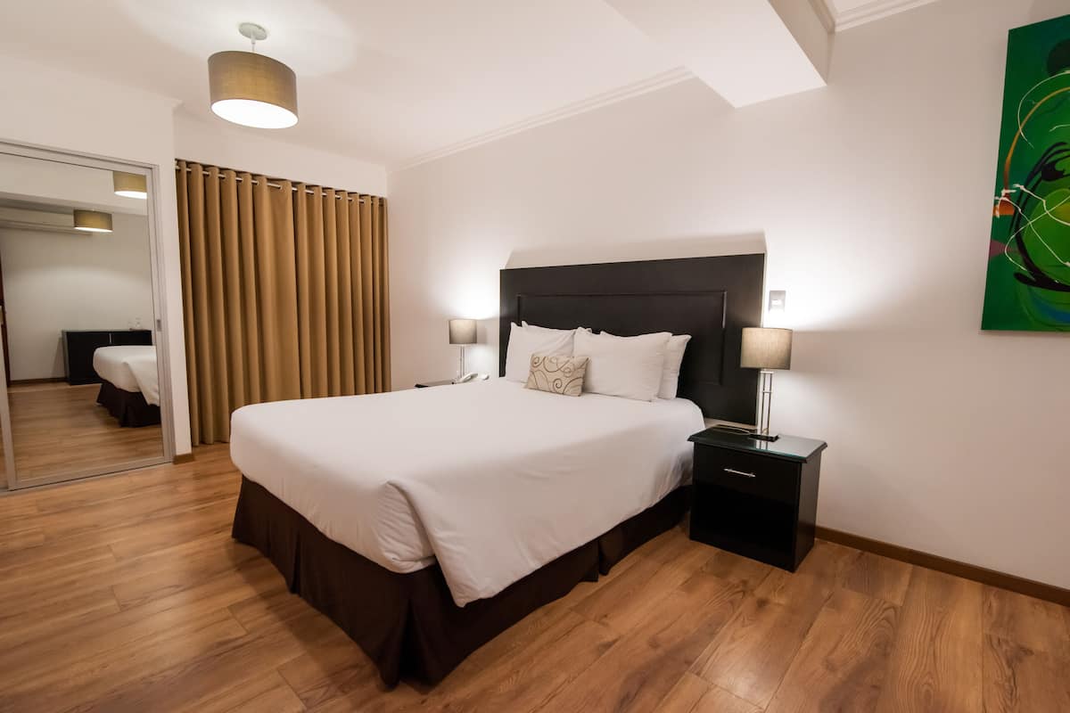 Une chambre d’hôtel avec un grand lit, une table de chevet avec une lampe, un miroir et un tableau abstrait vert accroché au mur.