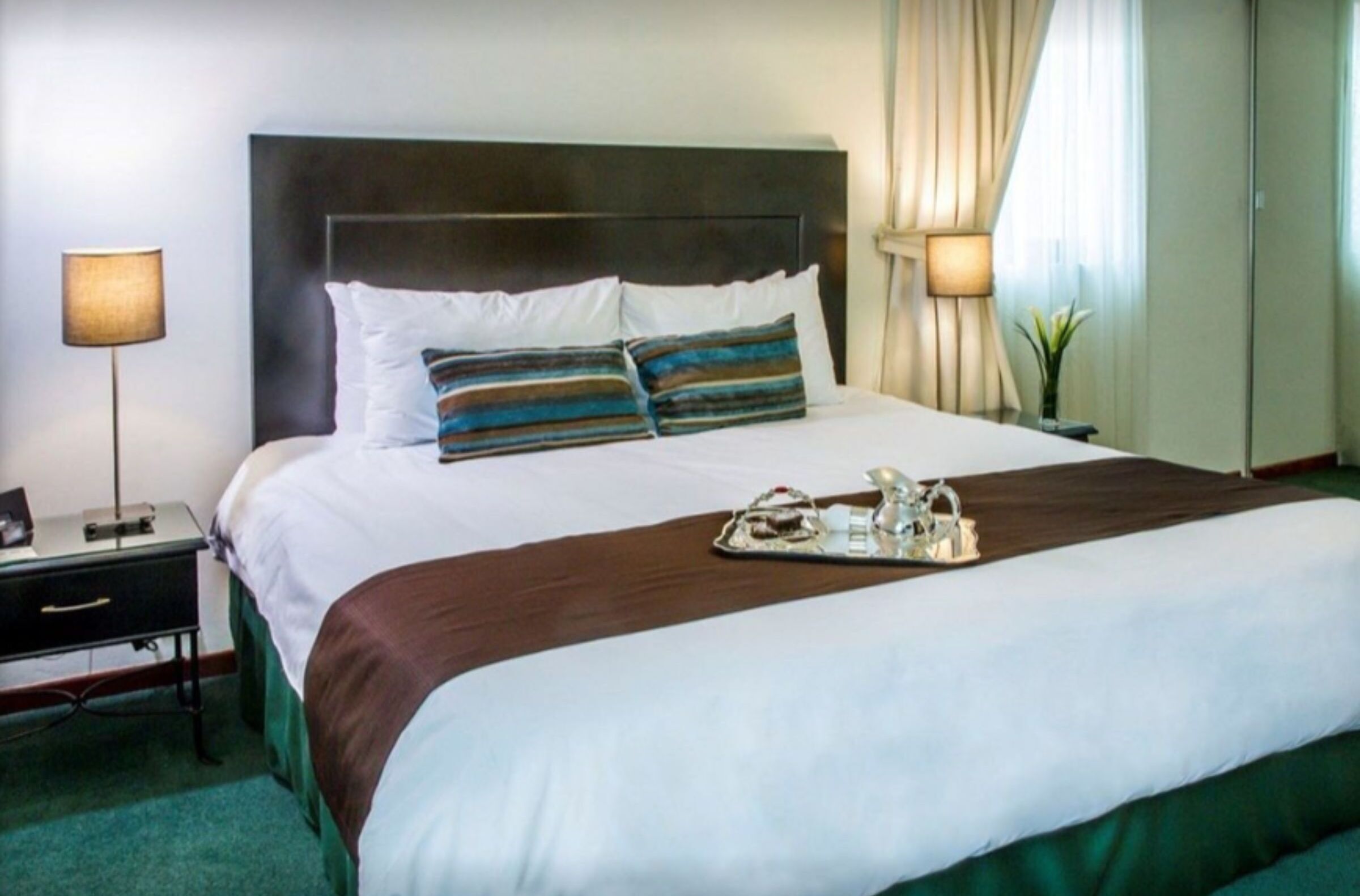 A hotel room with a large bed, two bedside lamps, a nightstand, and a tray with a teapot and cups.