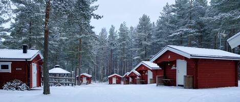 Une rangée de cabanes rouges aux bordures blanches, numérotées de 2 à 7, se dresse au cœur d’une forêt enneigée.