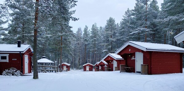 A row of red cabins with white trim, numbered from 2 to 7, in a snowy forest setting.