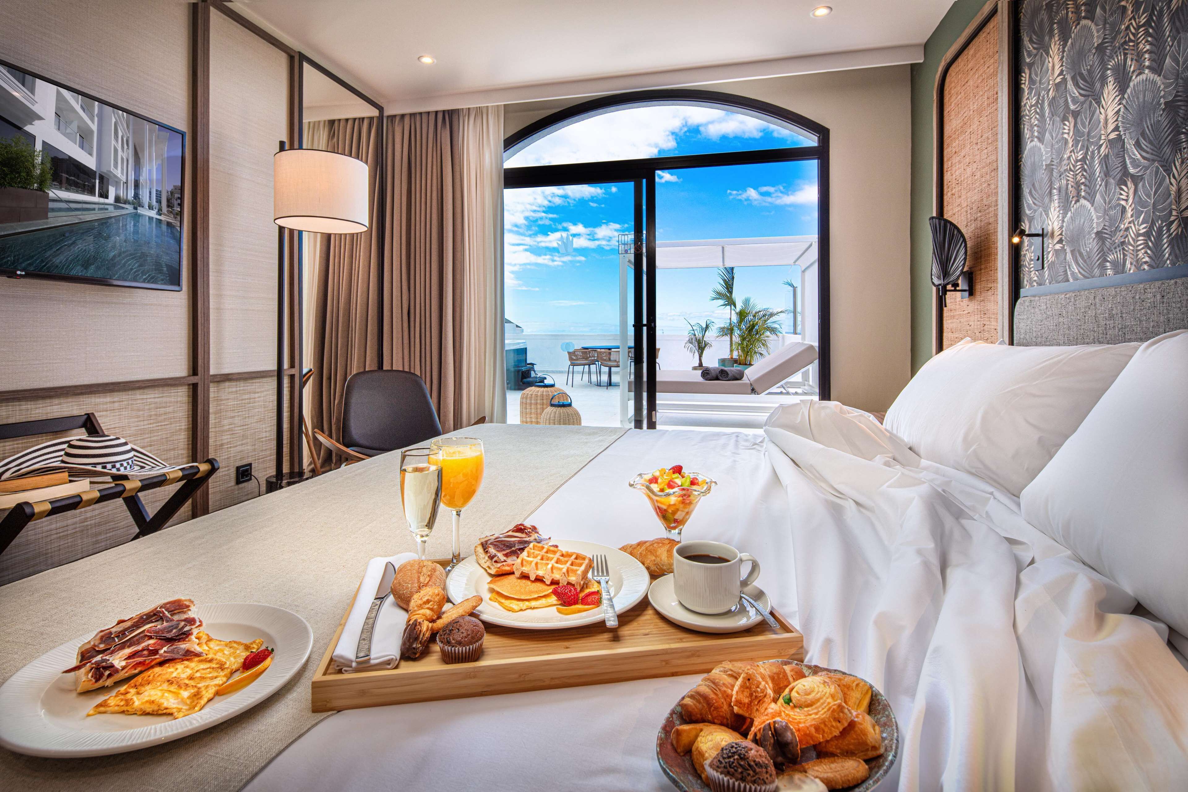 Habitación de hotel con bandeja de desayuno, cama y un ventanal con vista al océano.