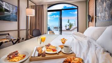 Habitación de hotel con bandeja de desayuno, cama y un ventanal con vista al océano.