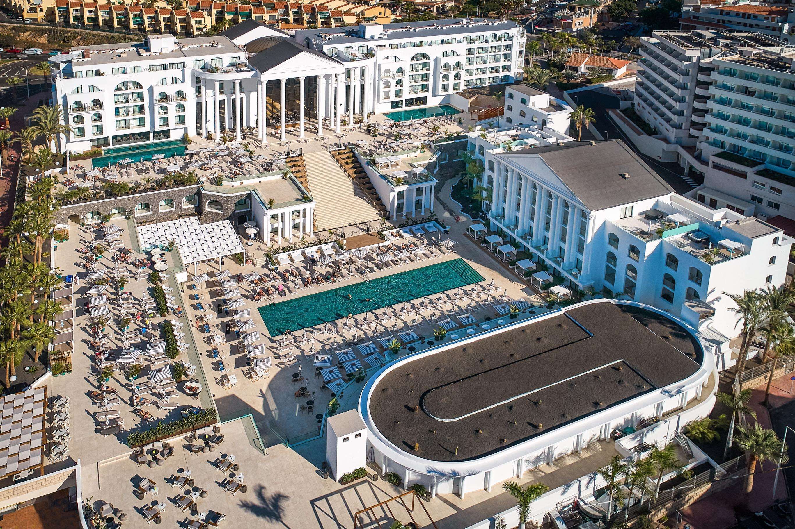 An aerial view of a hotel with multiple buildings, a central pool area, and numerous outdoor seating areas.