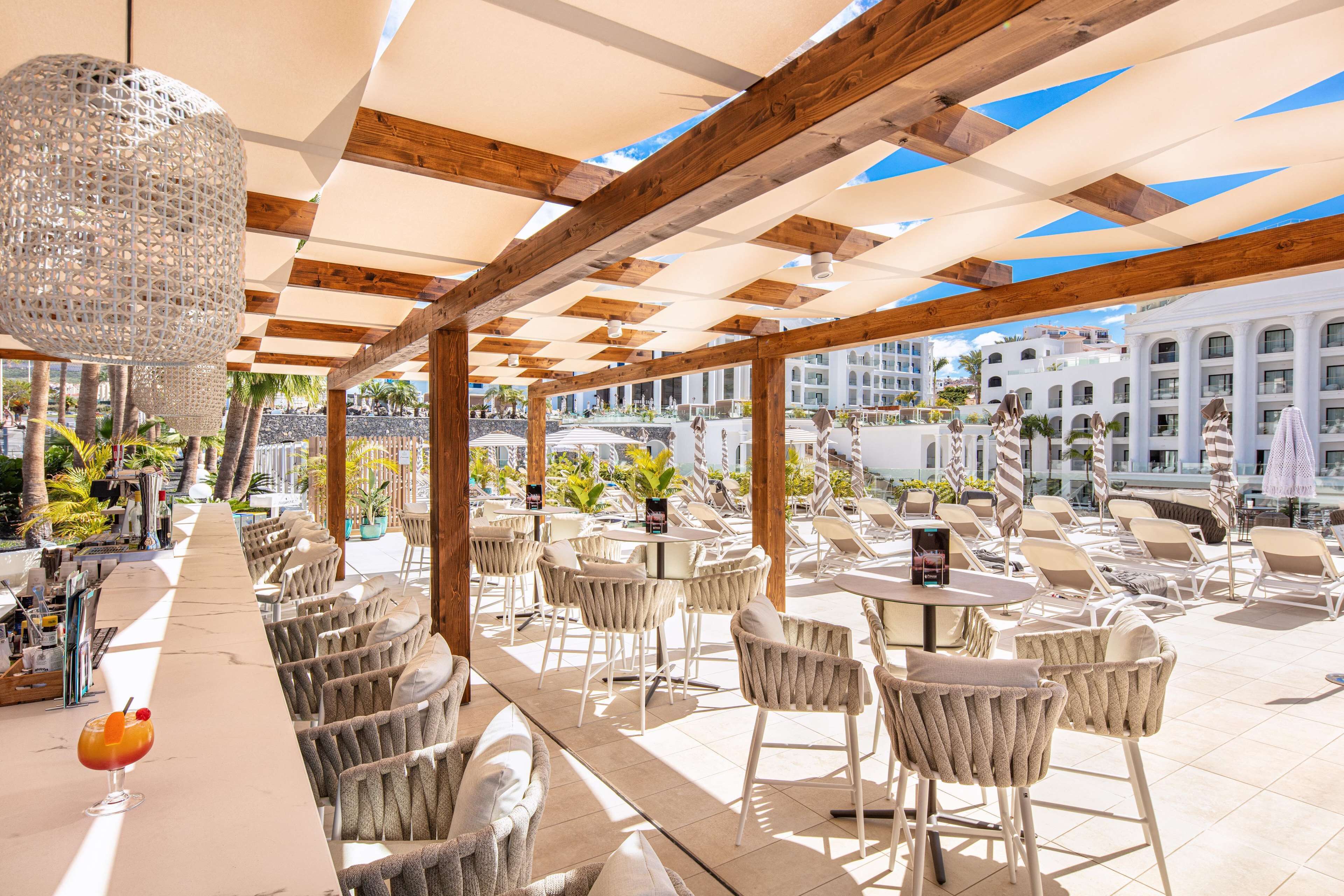 Un área de estar al aire libre iluminada por el sol, con vigas de madera y sillas blancas, con vistas a una piscina y edificios de la ciudad.