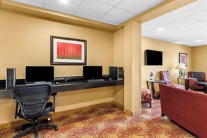 A hotel room with a desk, two laptops, a chair, a sofa, a TV, and a framed picture on the wall.