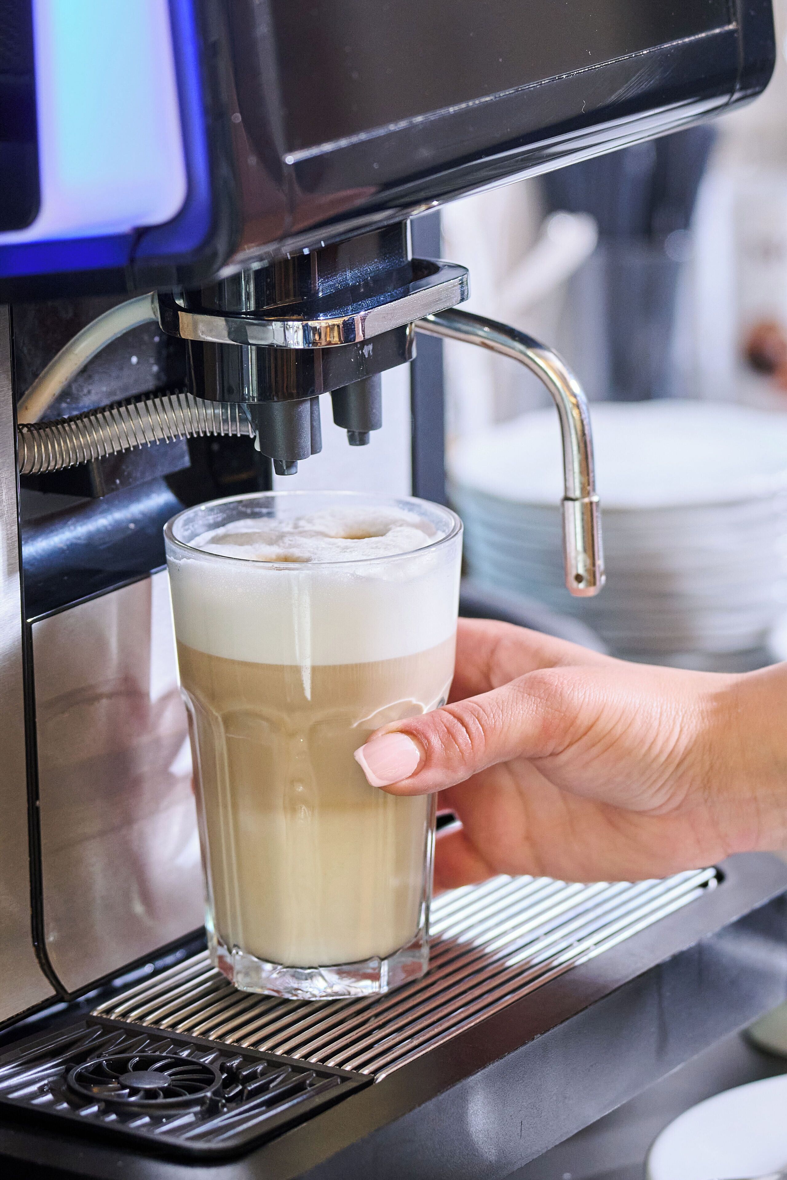 A hand holding a glass under a coffee machine dispensing a layered beverage.