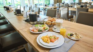 A breakfast buffet with various dishes, including eggs, ham, and croissants, in a bright dining area with large windows. - Hotel Heidehof Garni (Buedelsdorf)