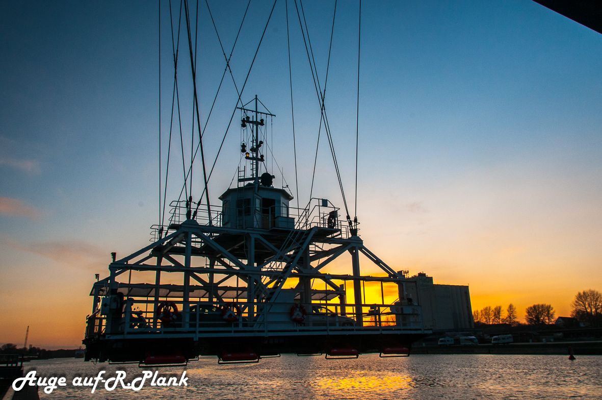 A ship with a crane and a cabin structure, docked at a port during sunset.