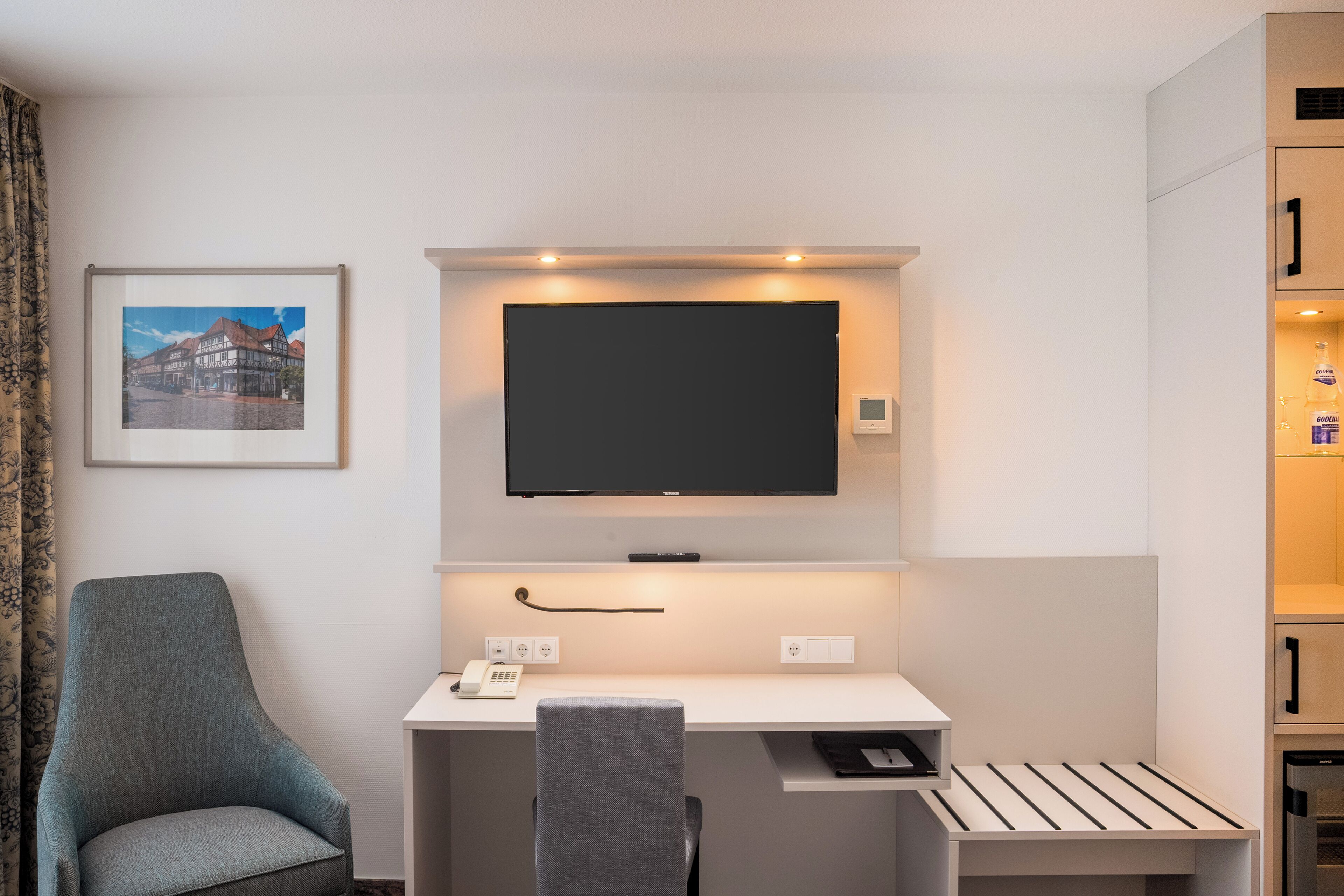Ein Hotelzimmer mit Schreibtisch, Fernseher darüber, einem Sessel und einem gerahmten Bild an der Wand.