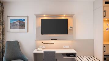 Ein Hotelzimmer mit Schreibtisch, Fernseher darüber, einem Sessel und einem gerahmten Bild an der Wand.