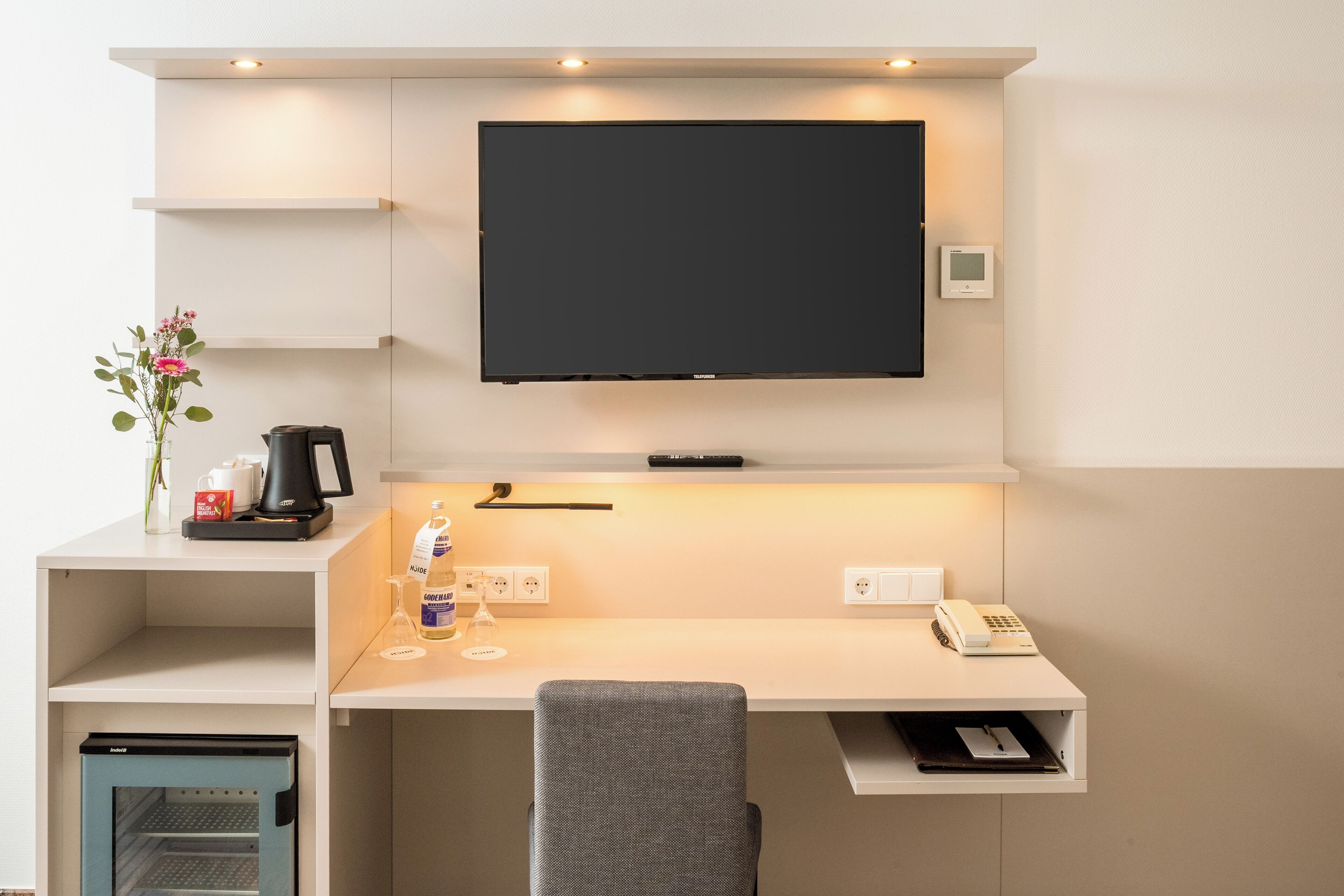 A modern hotel room with a mounted TV, a desk with a chair, a mini-fridge, a coffee maker, and a telephone.