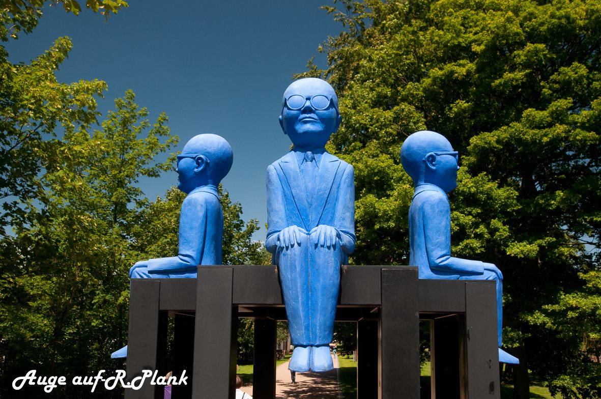 A sculpture of three figures seated on a platform with a clear sky and trees in the background.