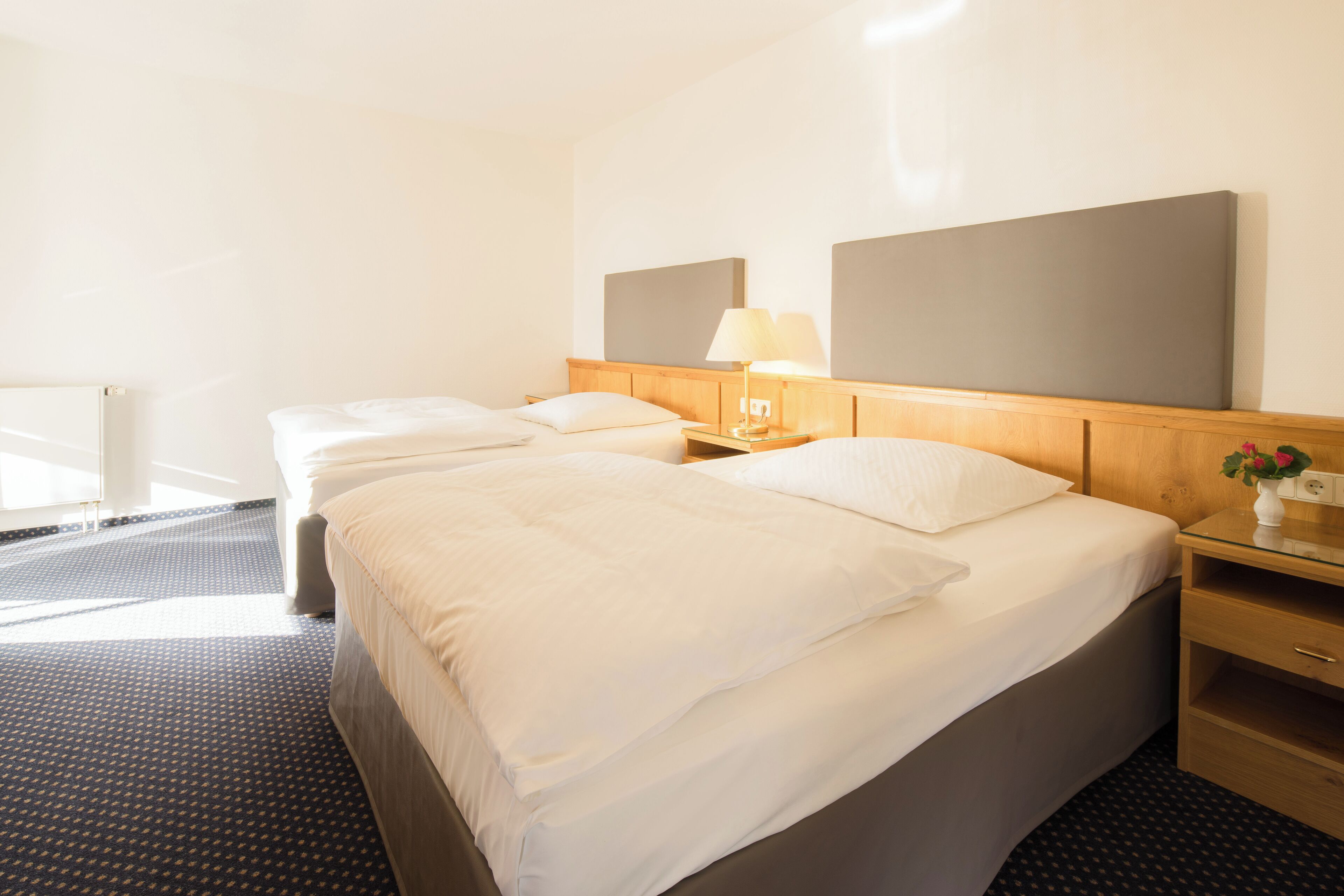A hotel room with two beds, a wooden headboard, bedside tables with lamps, and a small vase with flowers.