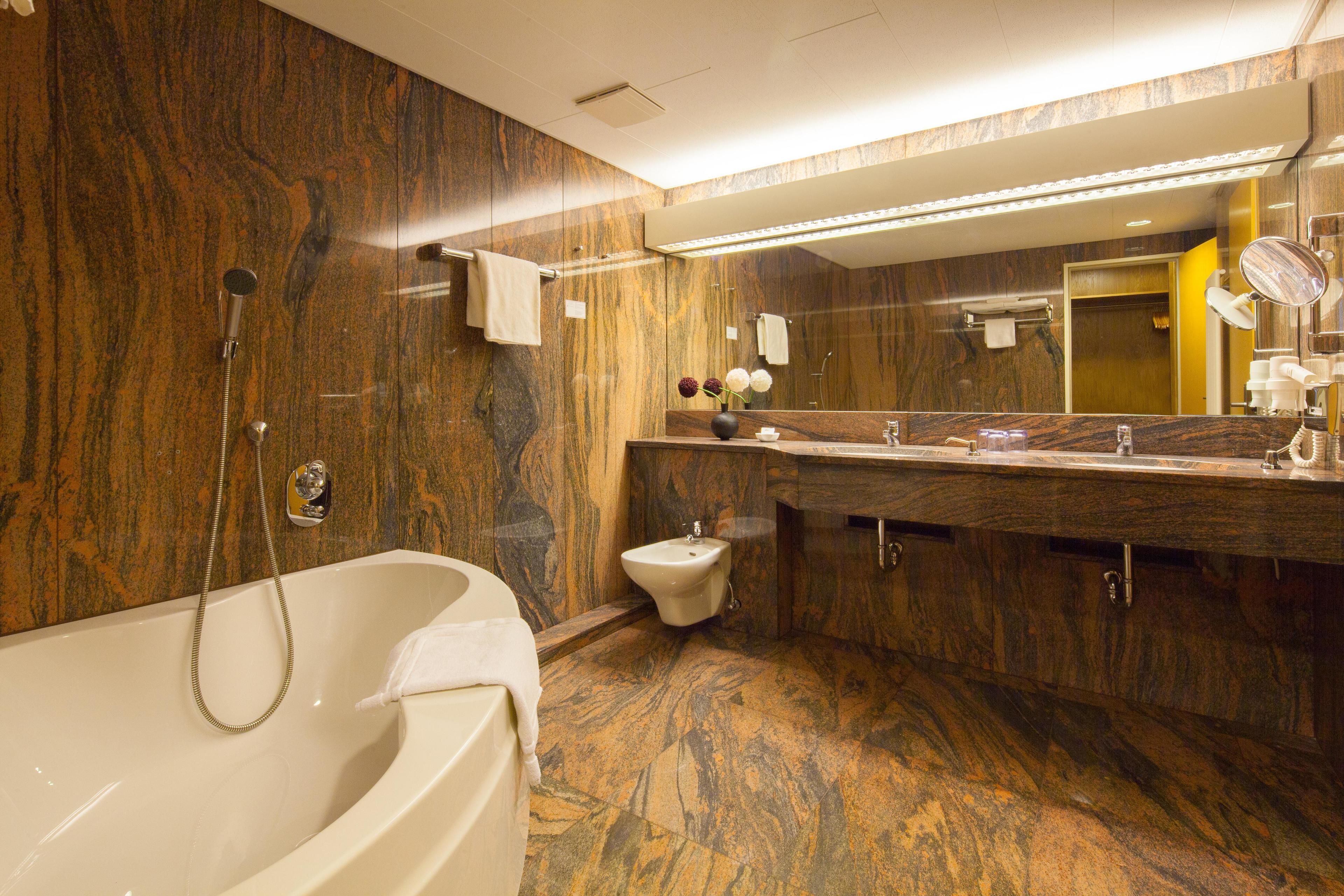A modern bathroom with a marble wall, a freestanding bathtub, a bidet, and a large mirror.