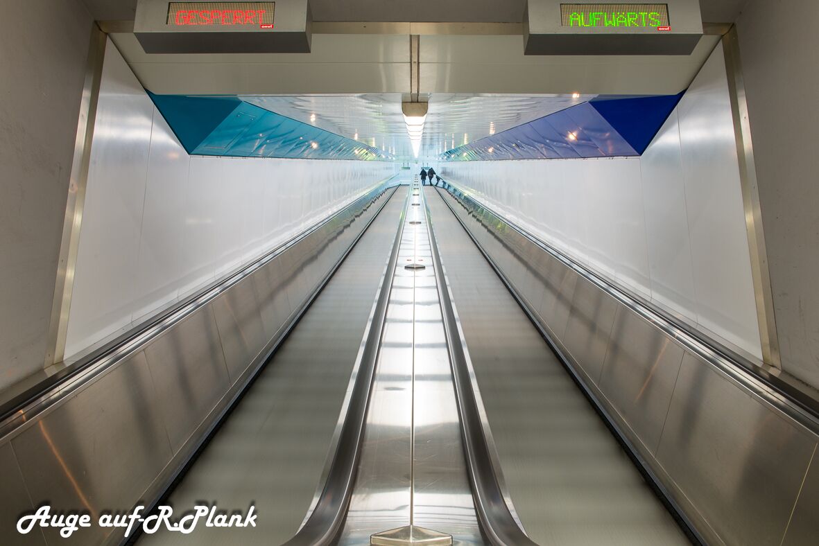 Una escalera mecánica en un espacio interior moderno y bien iluminado, con señalización digital arriba.
