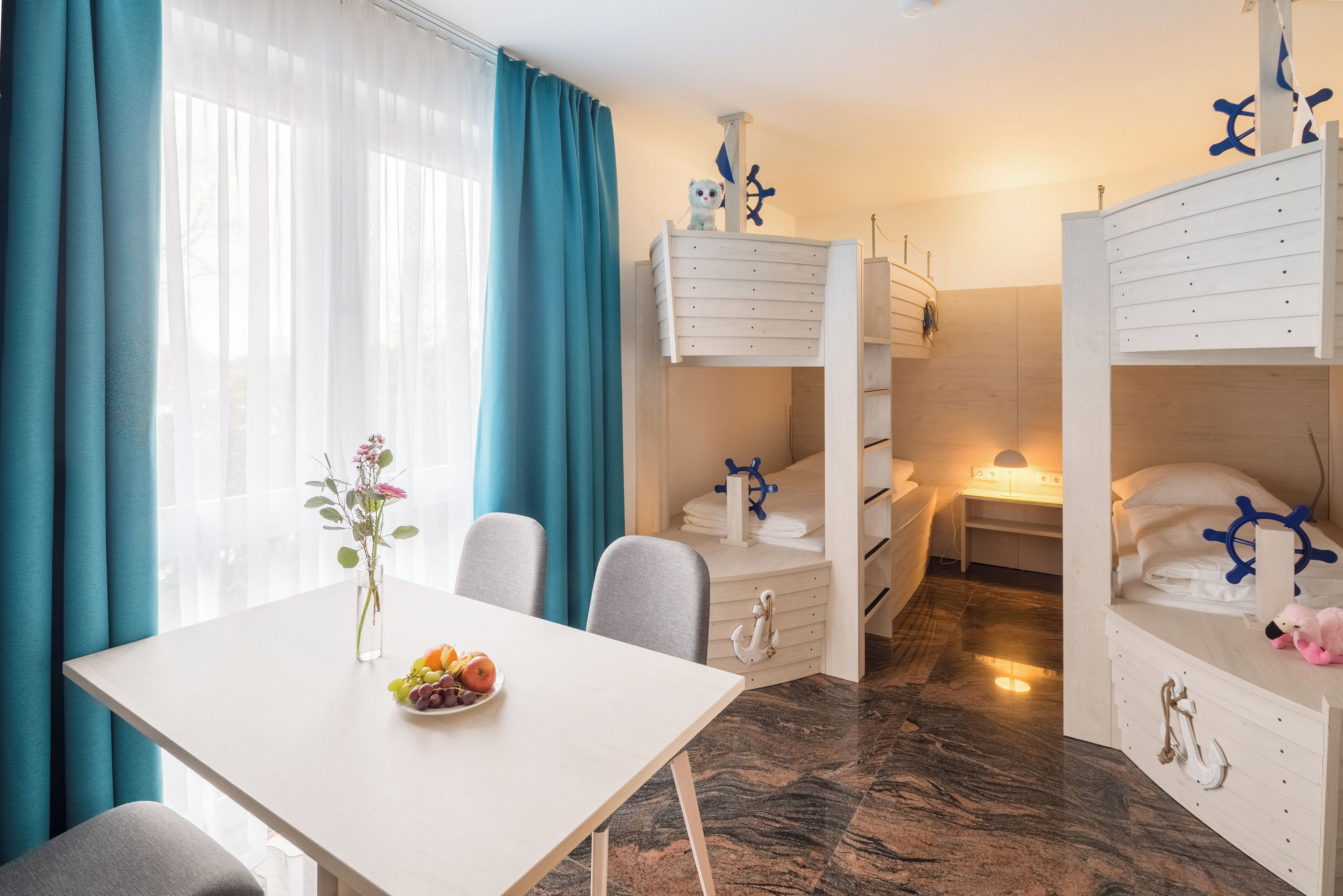Ett hotellrum med en våningssäng, ett skrivbord med en lampa, ett matbord med en vas med blommor och en utsikt genom ett fönster med gardiner.