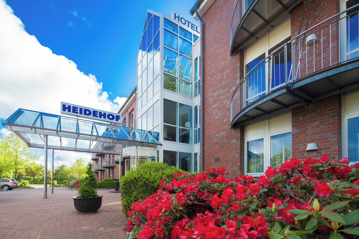 A hotel with a modern glass facade, a clear sky, and red flowers in the foreground.