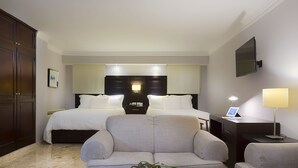 Ein Hotelzimmer mit einem Bett, zwei Nachttischen, einem Schreibtisch, einem Sessel und einer Couch. Ein Fernseher ist an der Wand befestigt.