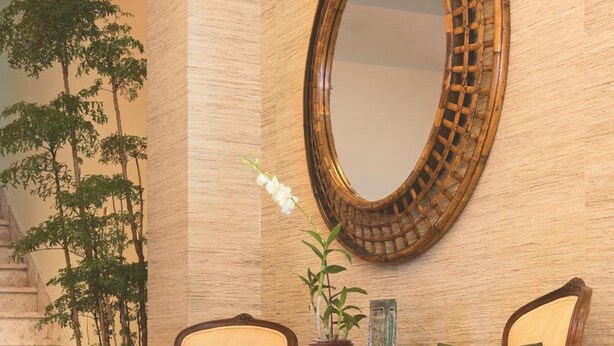 Eine Lobby mit einem großen Spiegel, zwei Holzstühlen mit Kissen, einem kleinen Tisch mit Pflanze und einer Treppe mit Grünpflanzen.