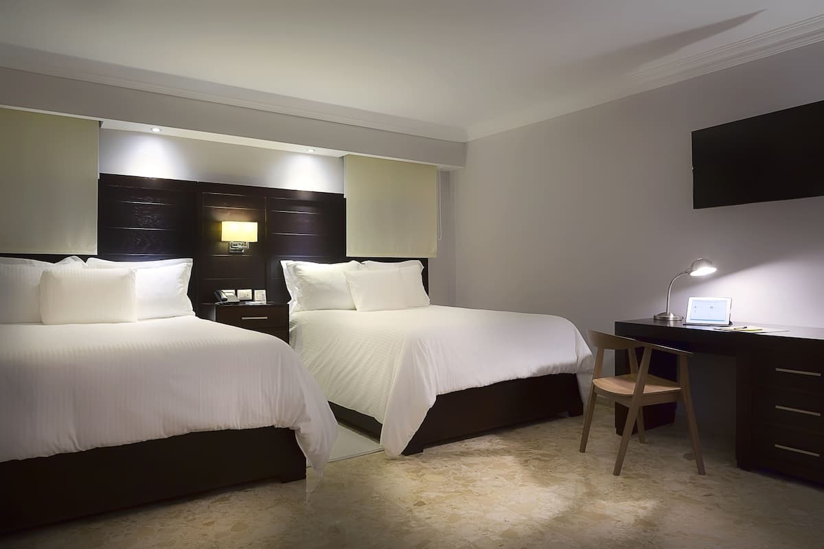 Une chambre d’hôtel avec deux lits, un bureau avec une chaise, un téléviseur à écran plat fixé au mur et une lampe sur le bureau.