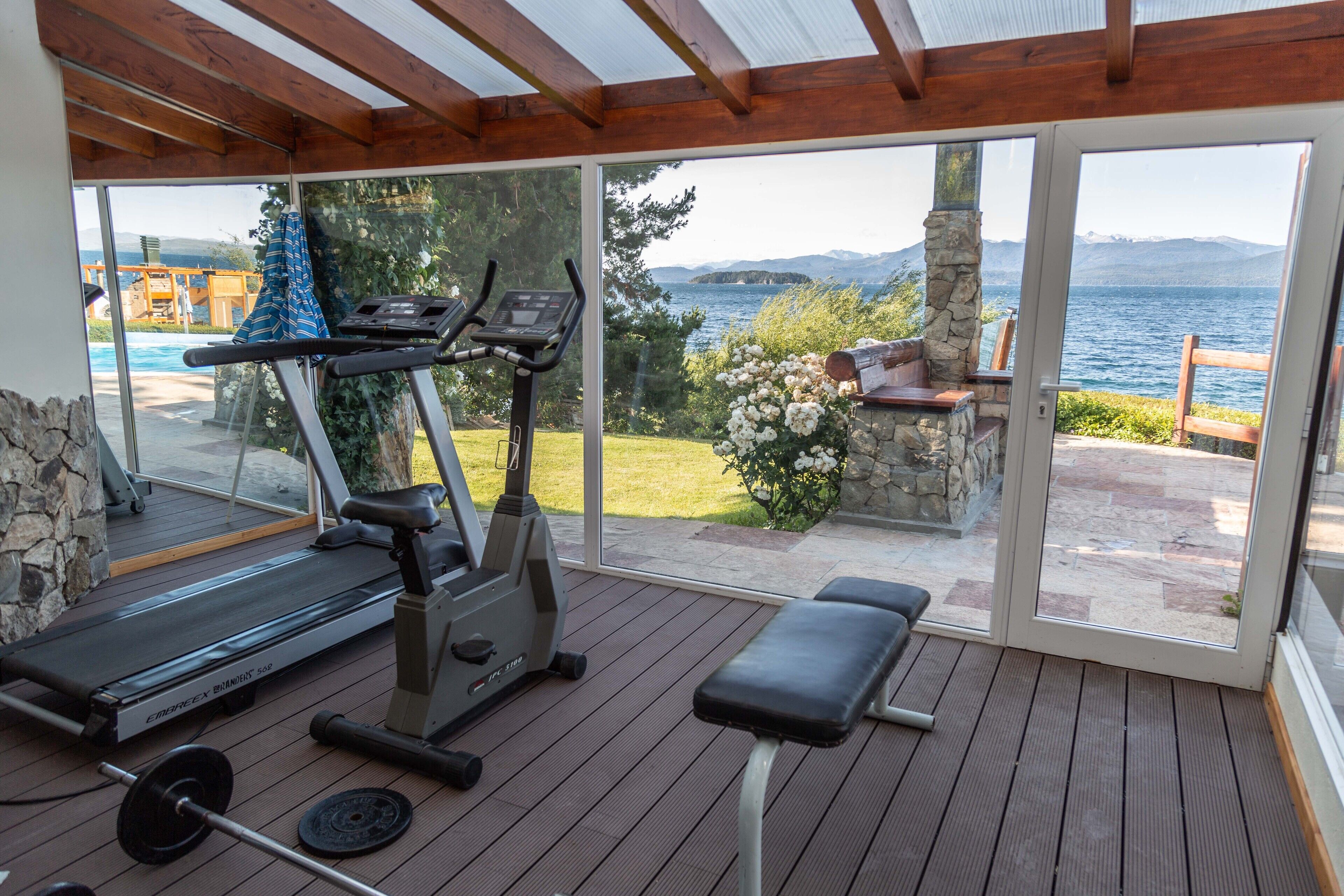 Zona de gimnasio al aire libre techada, con equipo de ejercicio, una banca y vistas a un lago y montañas.
