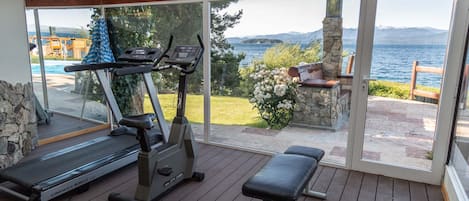 Zona de gimnasio al aire libre techada, con equipo de ejercicio, una banca y vistas a un lago y montañas.