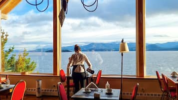 A dining area with a view of a lake and mountains, featuring a table with chairs and a person standing by the window.