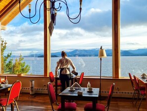Ein Essbereich mit Blick auf einen See und Berge, mit einem Tisch, Stühlen und einer Person, die am Fenster steht.