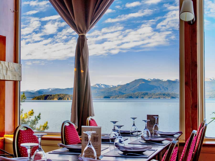A dining table set with red chairs, a candle, and a menu, overlooking a lake with mountains in the distance.