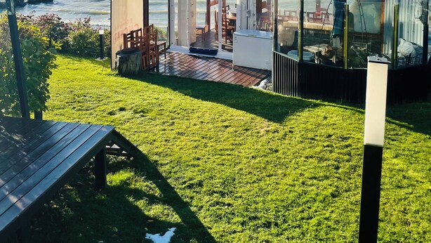 Un espacio al aire libre con paredes de vidrio que ofrece vistas al mar y a las montañas, muebles de madera y una pequeña mesa.