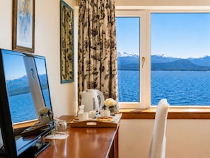 Ein Zimmer mit Schreibtisch, einer Kaffeemaschine, einem Tablett mit Speisen und einem Fenster mit Blick auf einen See und Berge.