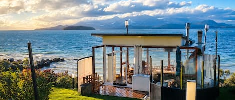 Estructura con paredes de vidrio, un deck y vistas a un cuerpo de agua y montañas.