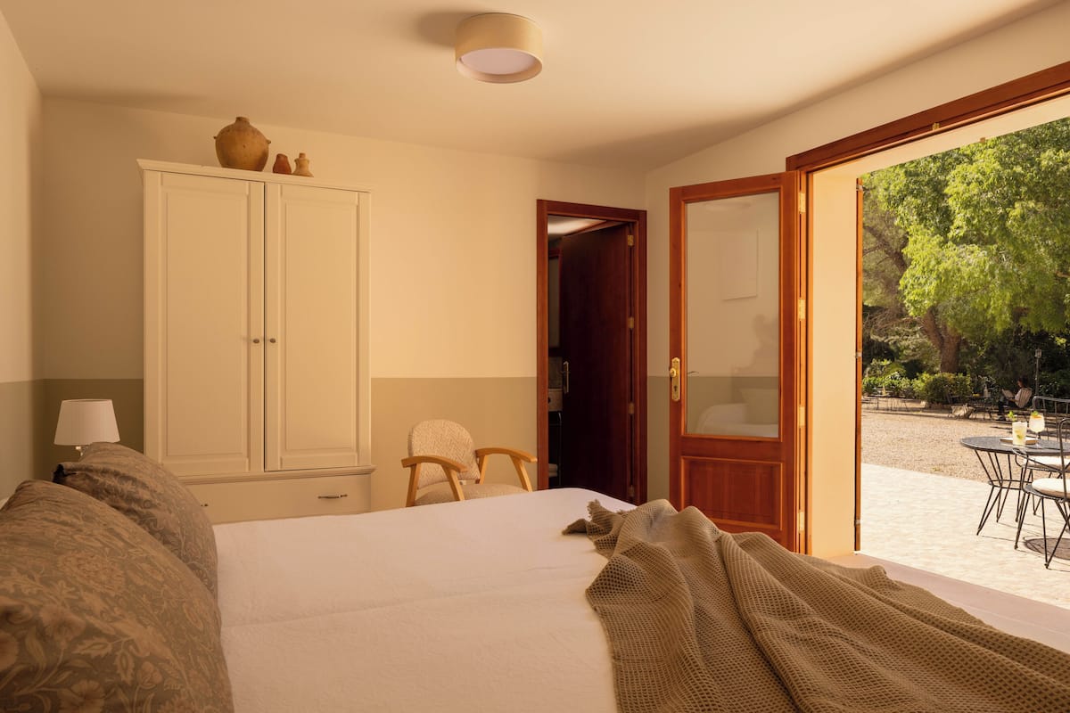 A bedroom with a bed, a wardrobe, a chair, and an open door leading to an outdoor area with a table and chairs.