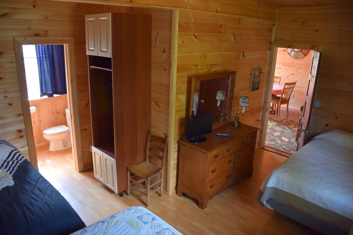 A wooden cabin room with a bed, a dresser, a chair, and a small bathroom with a toilet and sink.