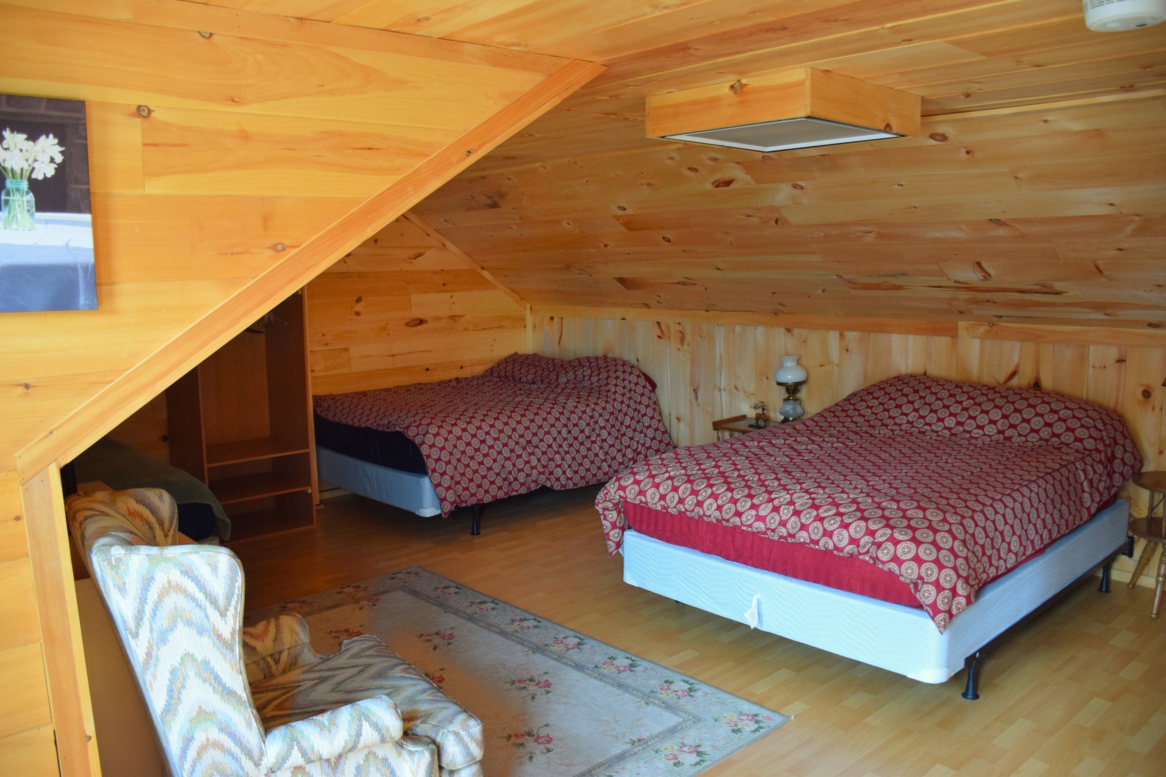A small, cozy attic bedroom with two beds, a chair, and a wooden ceiling.