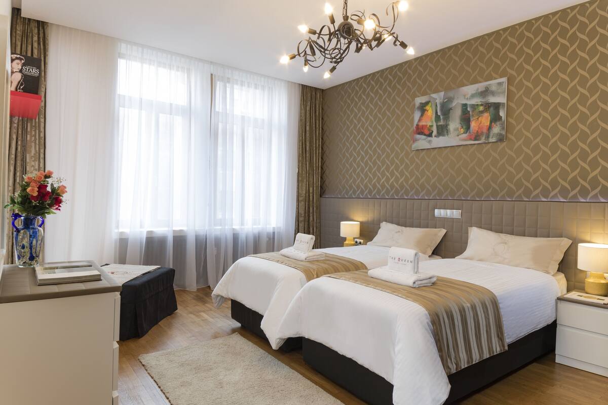Habitación de hotel con dos camas, un escritorio, una silla, un florero con flores, una araña y un cuadro en la pared.