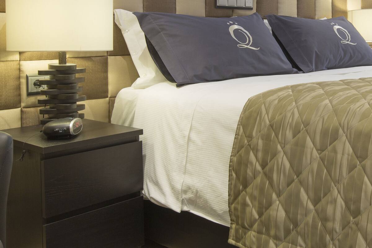 Una cama bien hecha con una cubierta acolchada, dos almohadas con un logotipo, una mesita de noche con una lámpara y un reloj fijado en la pared.
