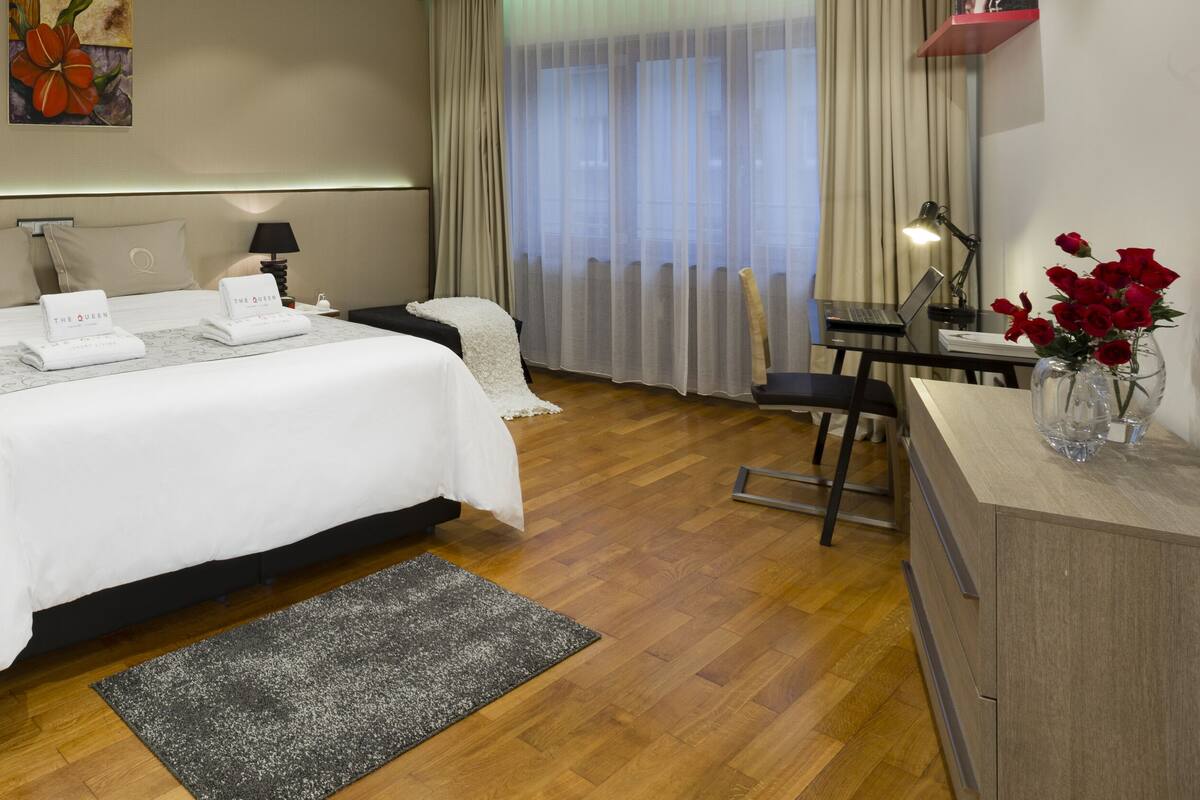 Una habitación de hotel con una cama grande, mesitas de noche con lámparas, un escritorio con una computadora portátil y un florero con flores.