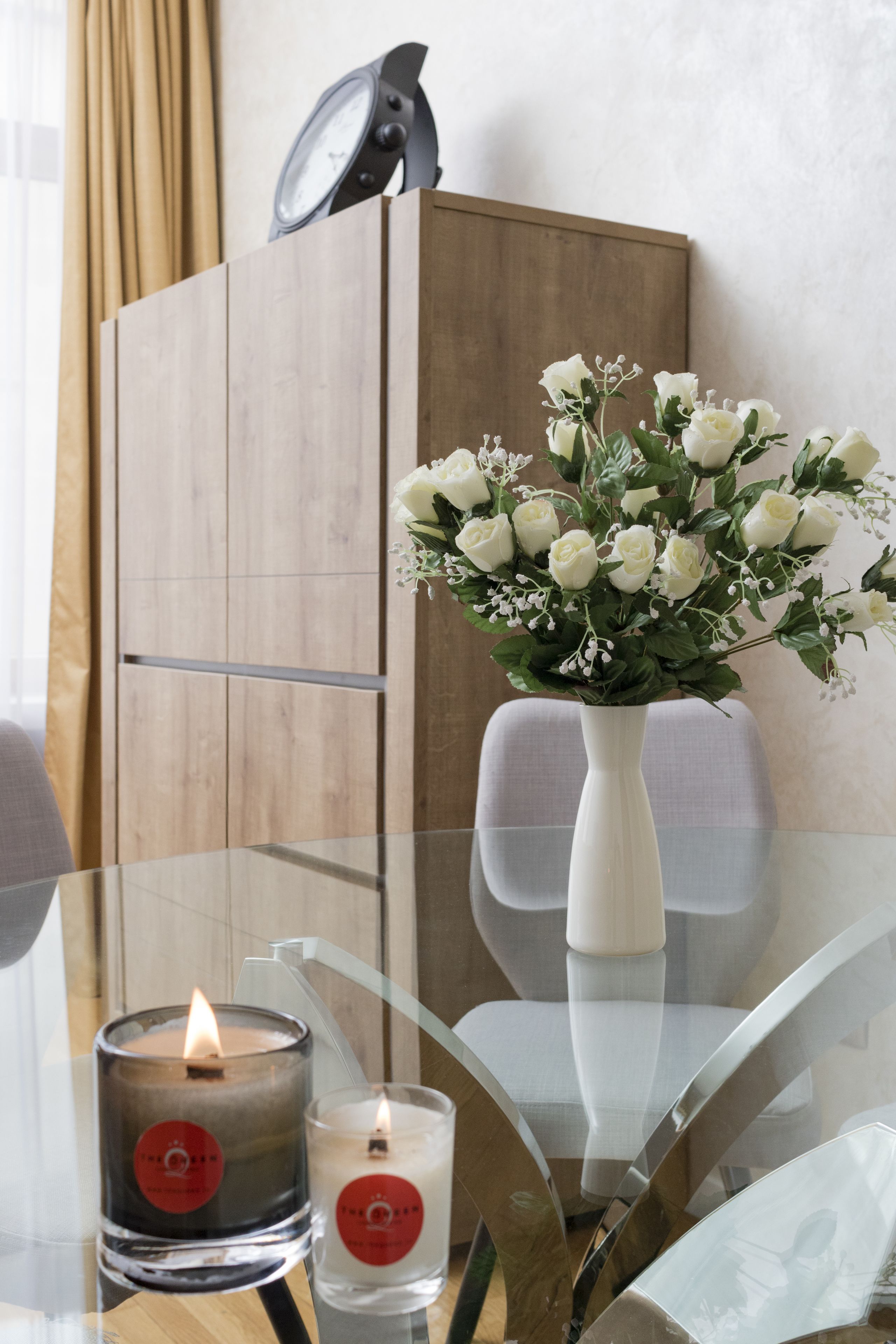 A glass table with two candles, a white vase with white flowers, and a wooden cabinet in the background.