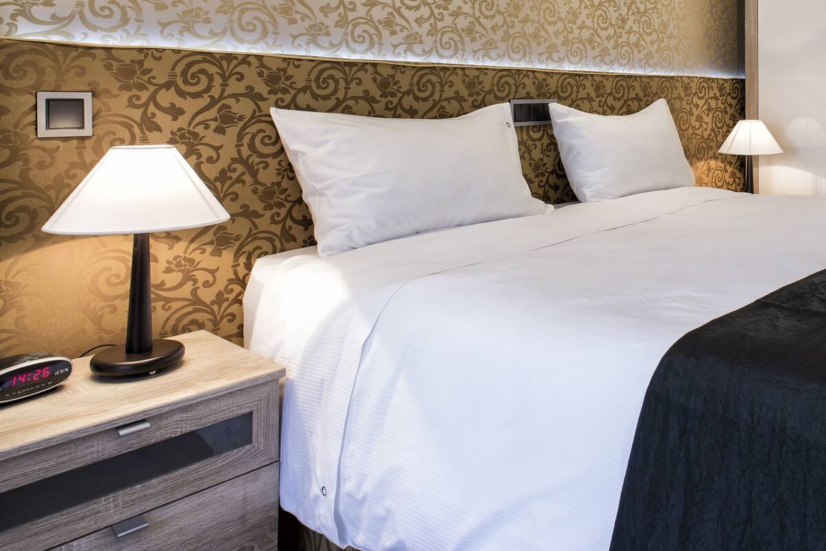 Una cama bien hecha con sábanas blancas, un plaid negro y toallas dobladas en el cabecero. Hay dos lámparas de noche en una mesita.