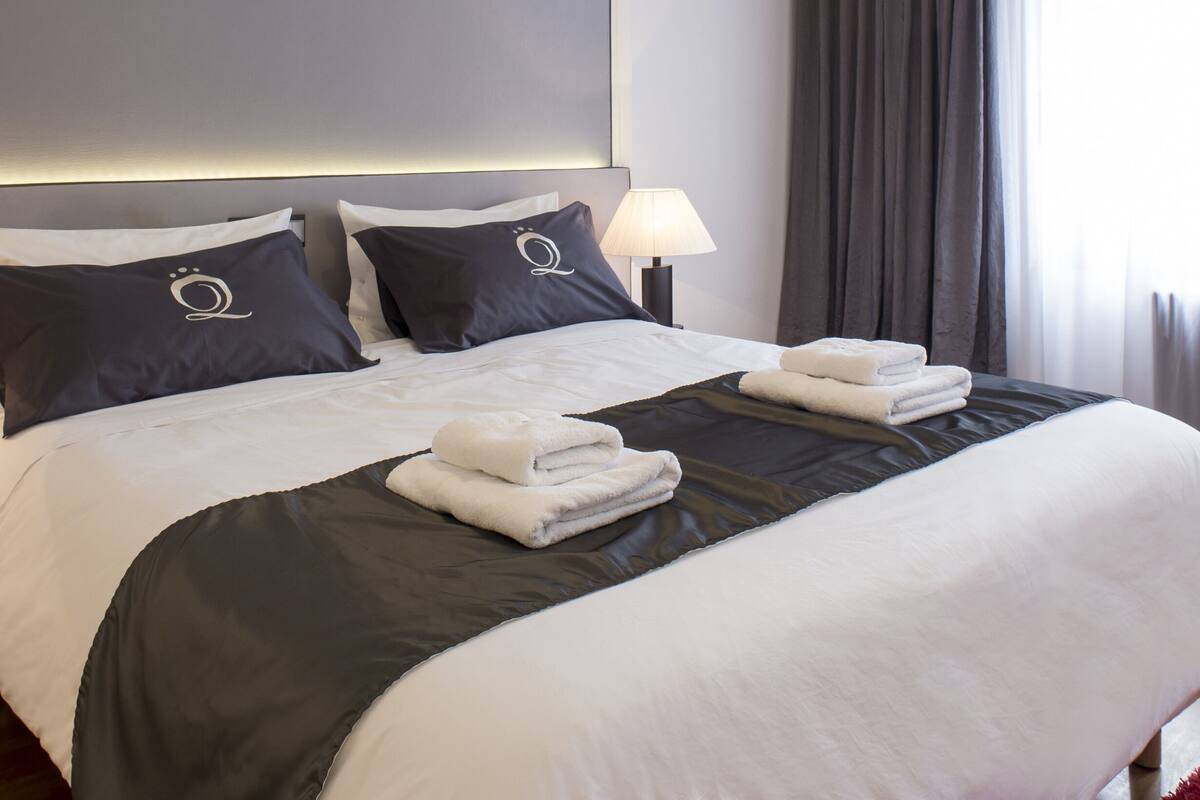 Una cama bien hecha con ropa de cama blanca y gris oscuro, dos lámparas de noche y una pila de toallas dobladas sobre la cama.