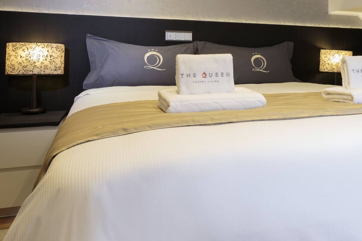 Una cama bien hecha con dos toallas, cada una con la inscripción 'THE QUEEN LUXURY LIVING', en una habitación de hotel moderna con cabecera y lámparas de noche oscuras.