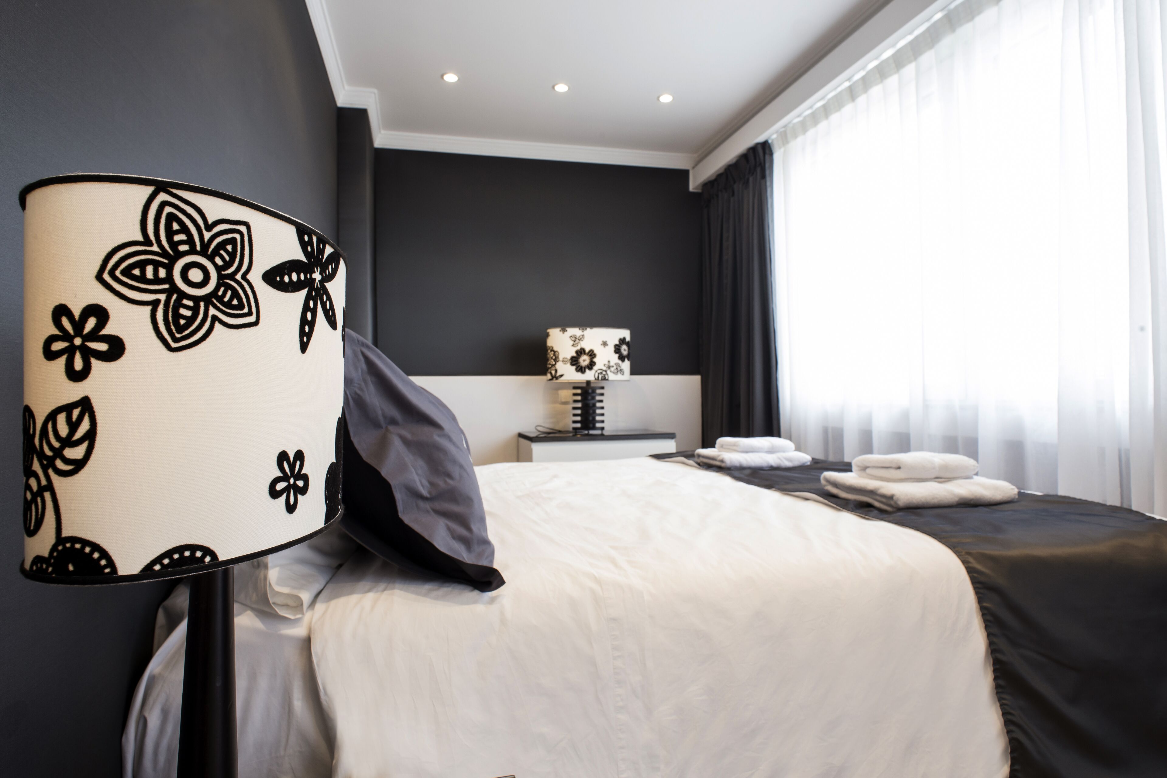 A modern hotel room with a large bed, a bedside table, a lamp, and a view of the outside through the window.