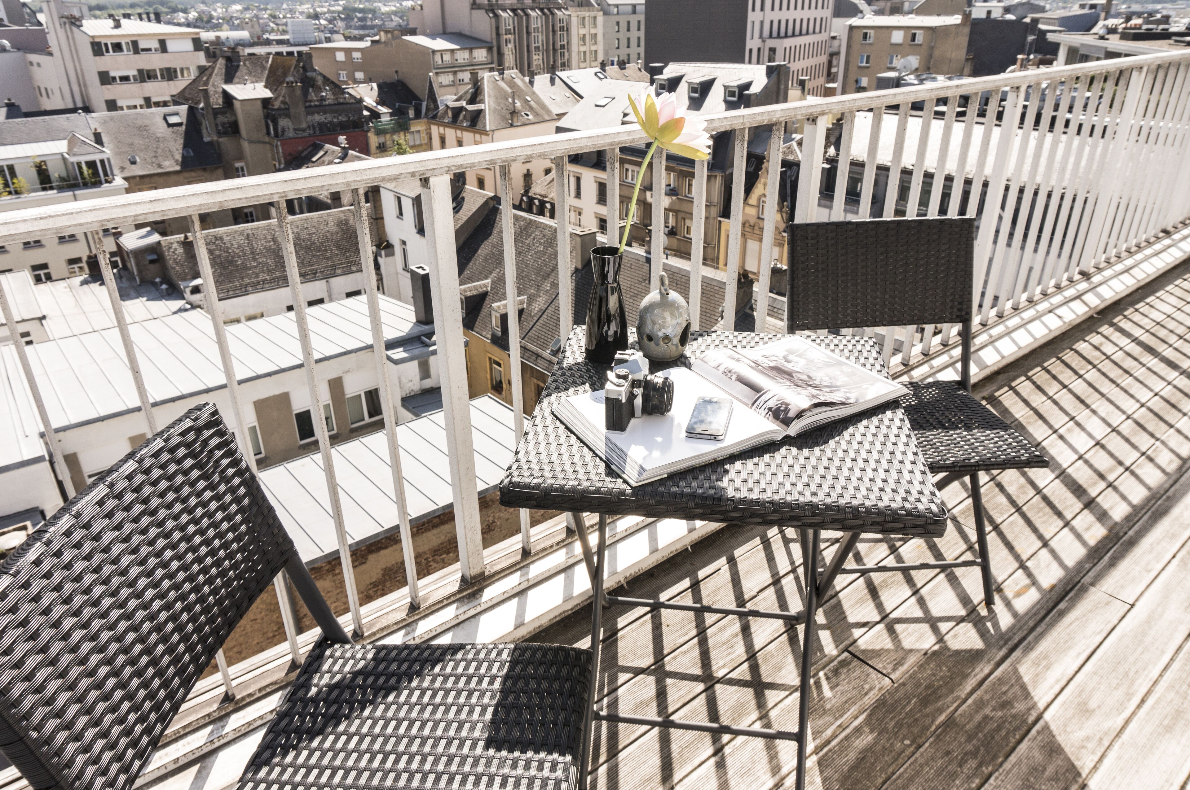 Une terrasse sur le toit, avec des fauteuils en osier et une table, un vase avec une fleur, et une vue sur la ville.