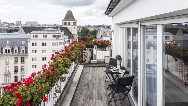 A balcony with a wooden floor, a white railing, and red flowers. There is a table with chairs and a window. - The Queen Luxury Apartments - Villa Fiorita (Luxembourg City)