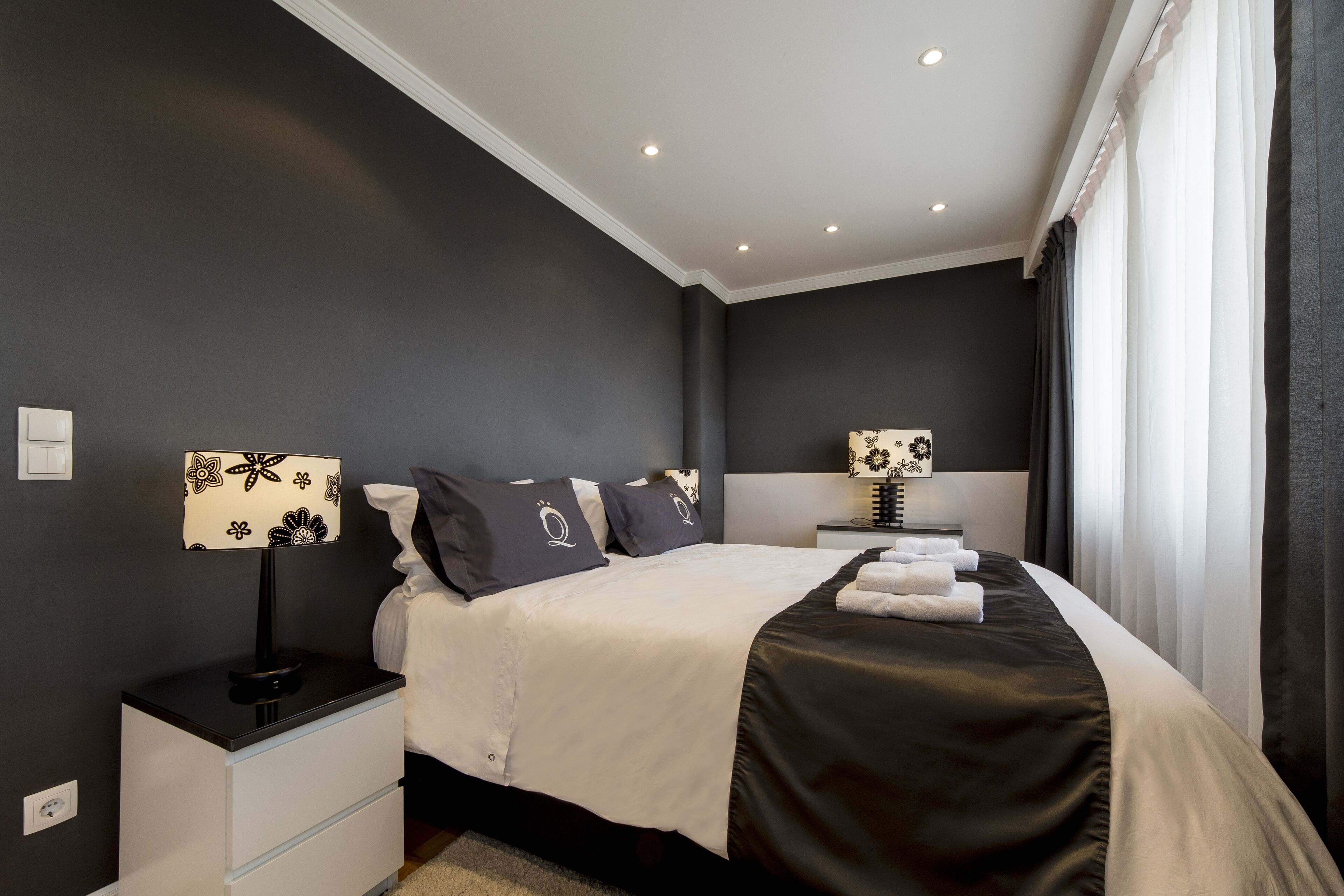 Une chambre moderne avec un mur foncé, un lit blanc, deux tables de chevet, une lampe et une fenêtre avec des rideaux.